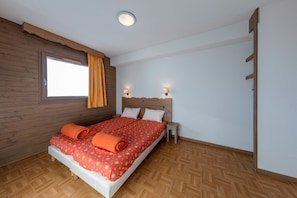 A bedroom with a wooden headboard, a bed with a red patterned bedspread, two bedside lamps, and a window with curtains. - Noemys L'Ecrin des Neiges (Chamrousse)