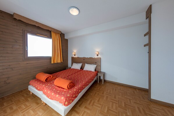 Un dormitorio con cabecera de madera, una cama con colcha estampada roja, dos lámparas de noche y un ventanal con cortinas.