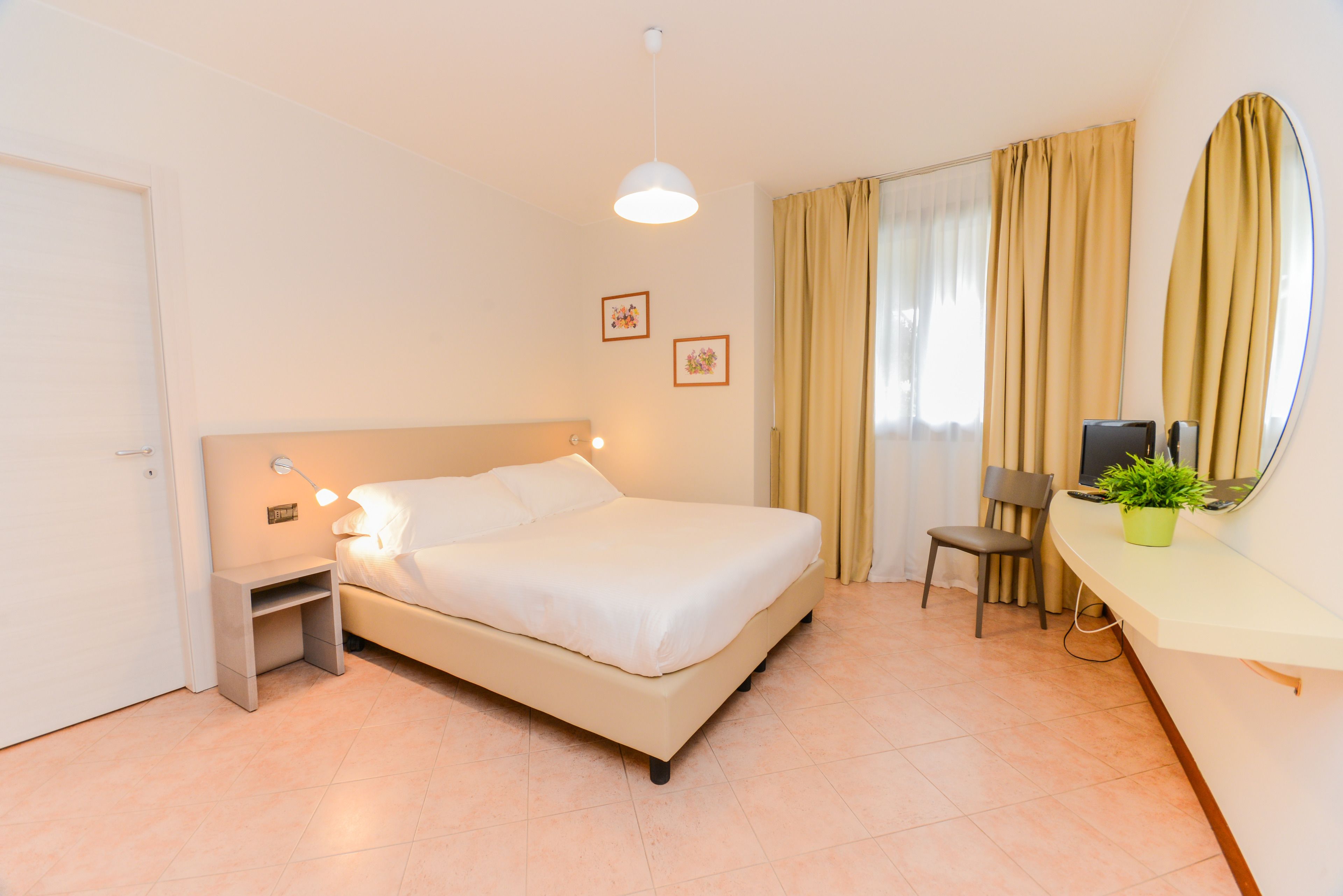 A hotel room with a bed, bedside table, chair, desk, and a potted plant.