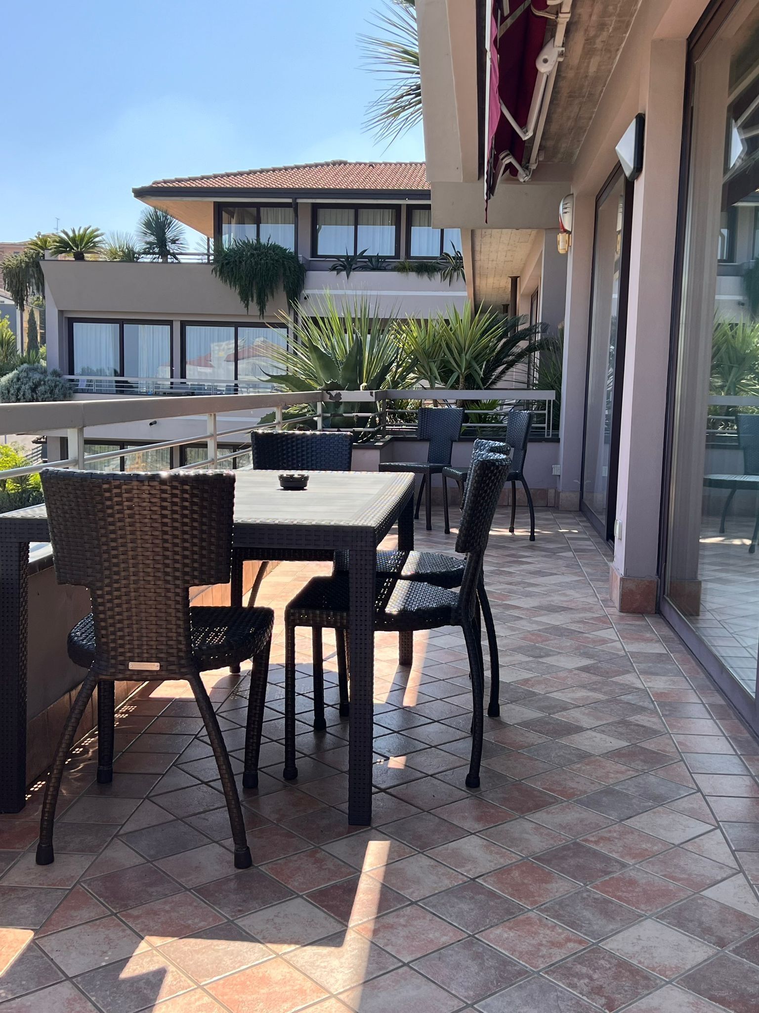 Ein Außenbereich mit Terrasse, einem Tisch, Stühlen, einem Fliesenboden und Blick auf ein Gebäude mit einem Ziegeldach.