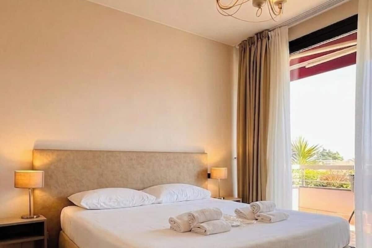 Ein Hotelzimmer mit einem großen Bett, zwei Nachttischlampen, einer Kristallleuchtre und Blick ins Freie.