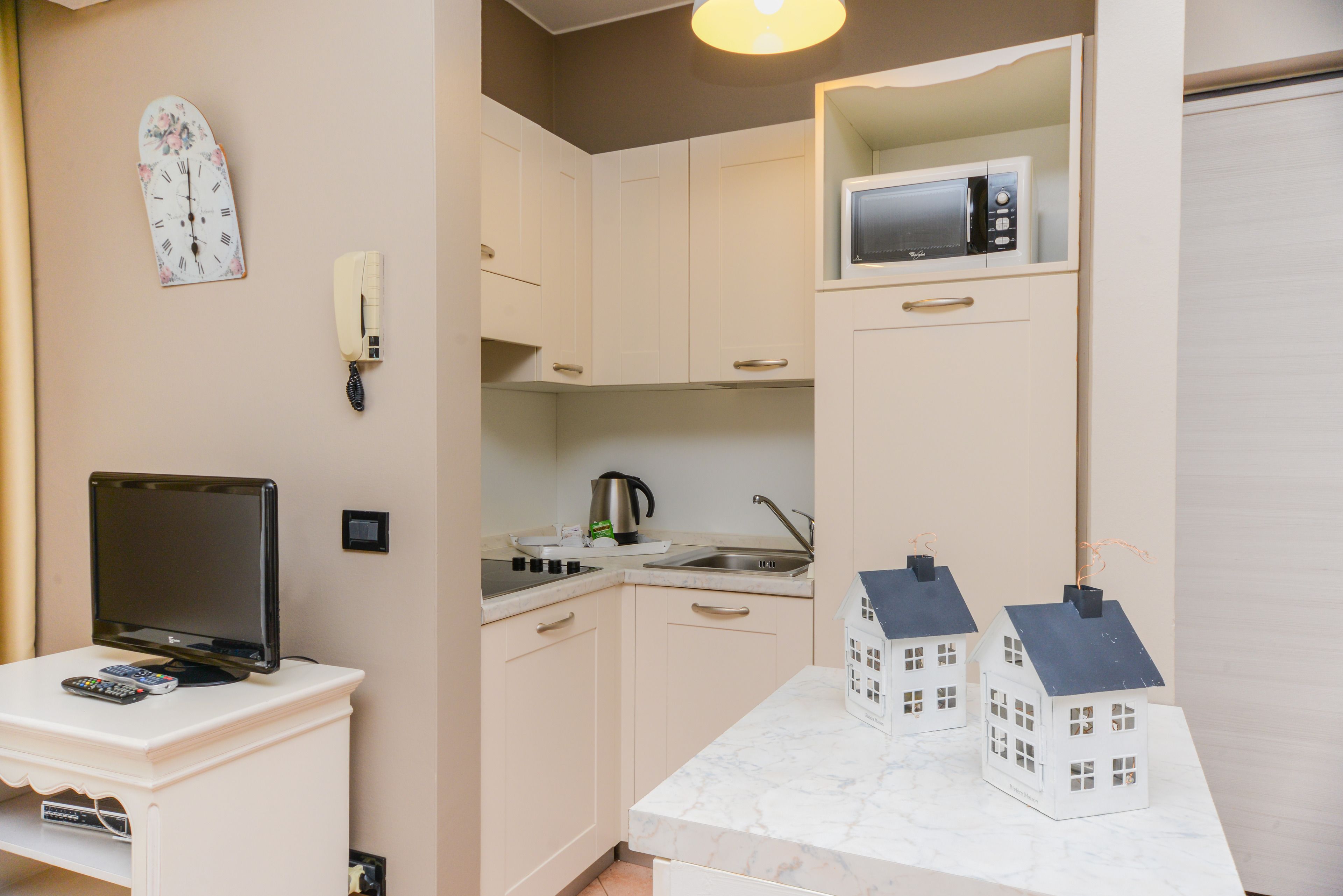 Een compacte keuken met een magnetron, waterkoker en twee kleine decoraties in de vorm van huisjes op het aanrecht.