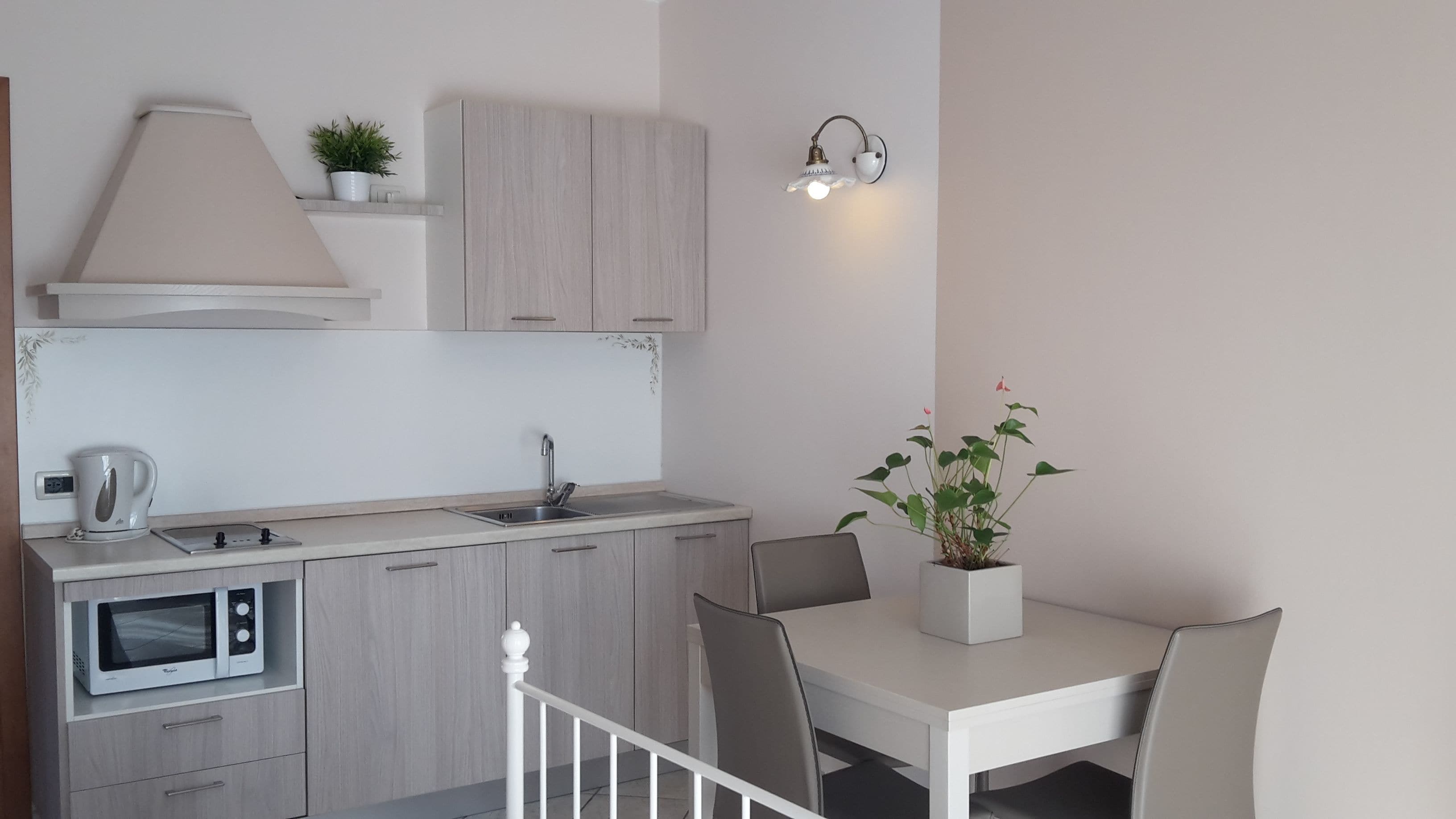 A modern kitchen with a microwave, kettle, sink, and cabinets. There is a dining table with chairs and a potted plant.