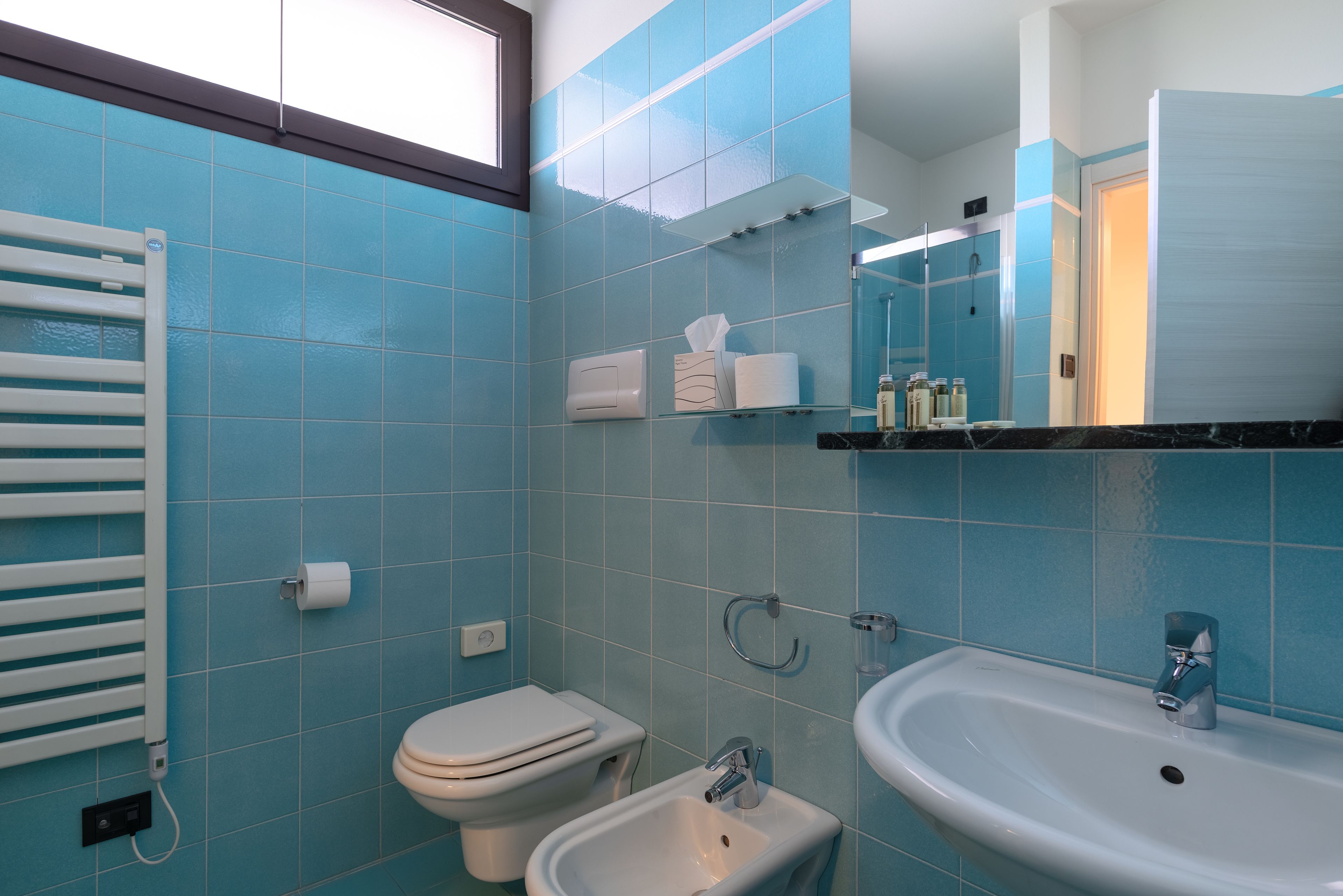 A bathroom with blue tiles, a toilet, a bidet, a sink, and a towel dispenser.