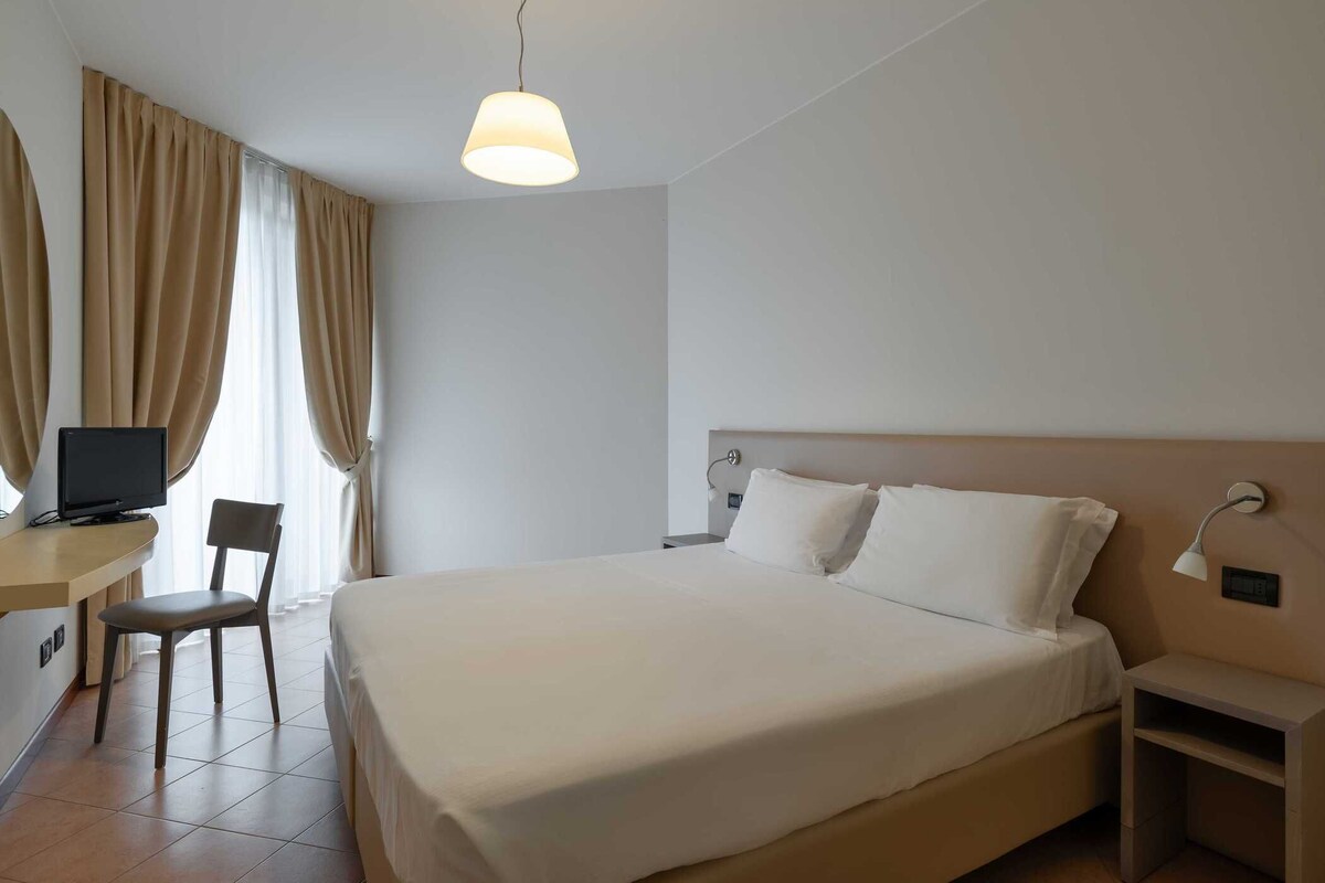 A hotel room with a large bed, a desk with a chair, a television, and a bedside table with a lamp.