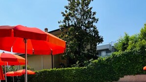 A poolside area with red lounge chairs and umbrellas, a clear blue pool, and a building in the background. - Il Sogno Apartments (Desenzano del Garda)