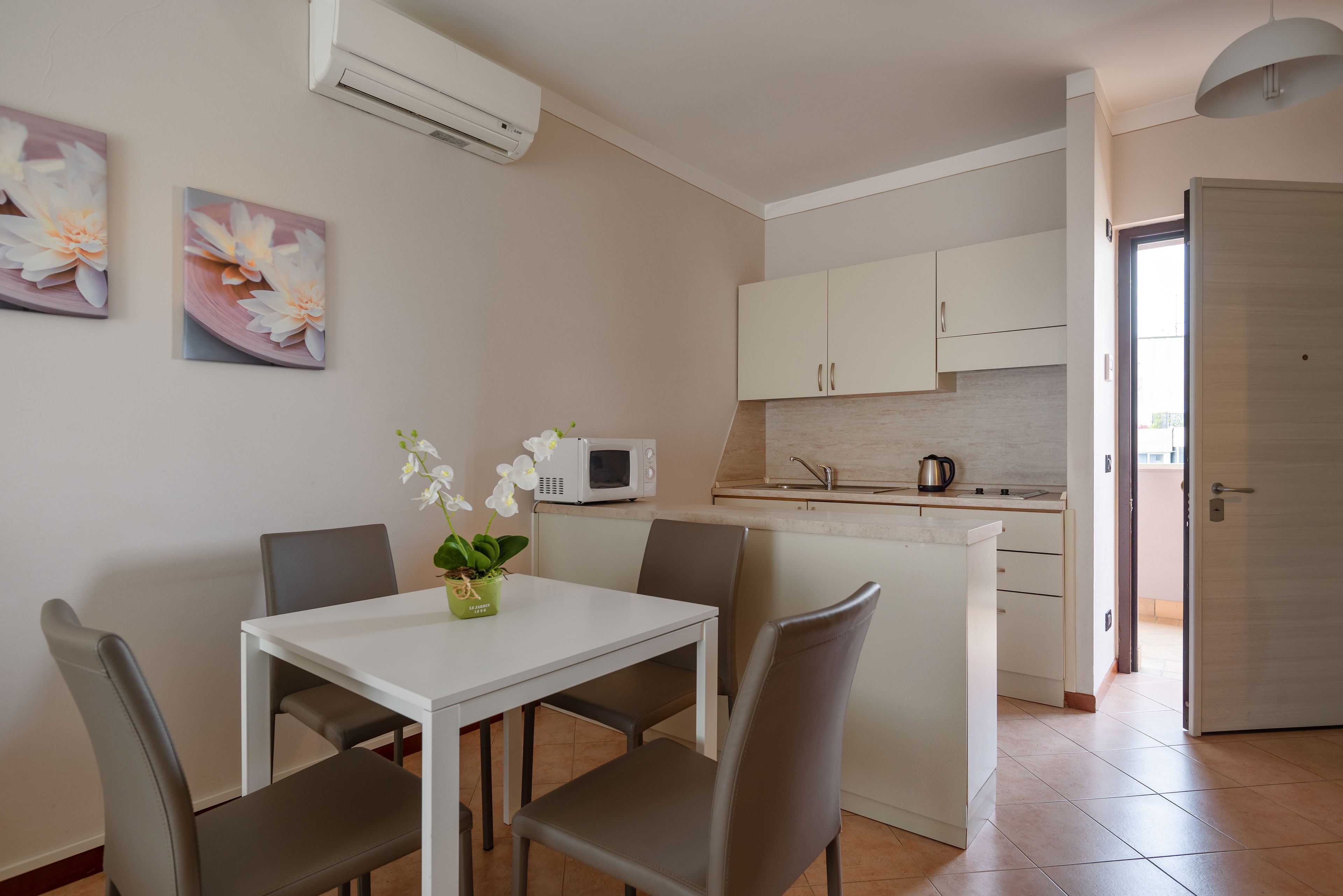 A modern kitchen with white cabinets and a microwave, a dining area with a white table and chairs, and a door leading to another room.