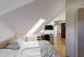 Quarto de hotel moderno com teto inclinado, cama com roupa de cama branca, escrivaninha com computador e TV de tela plana na parede.
