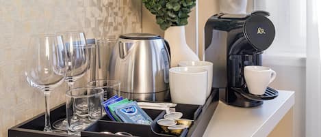 Un plateau avec une machine à café, une bouilloire, un vase contenant des plantes vertes, un présentoir de capsules de café, une tasse et des verres à vin.