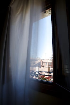 A window with a curtain partially drawn, revealing a balcony with a railing and some flowers.