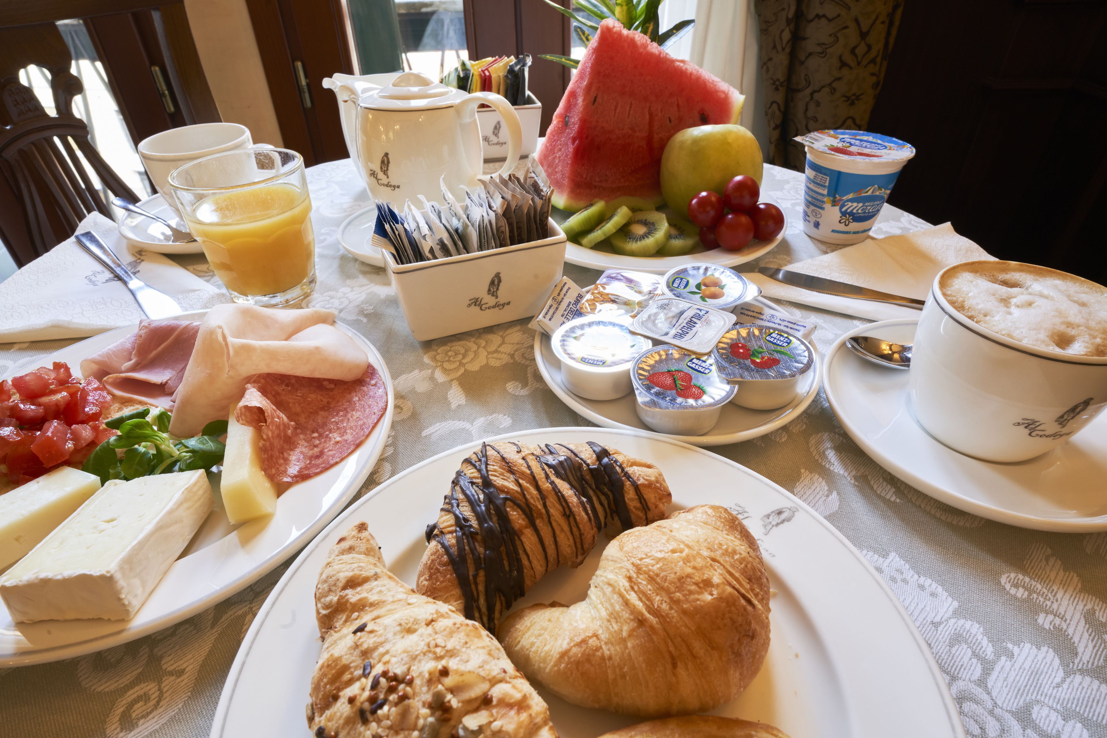 Mesa de café da manhã com diversos alimentos, incluindo croissants, queijo, presunto e frutas.
