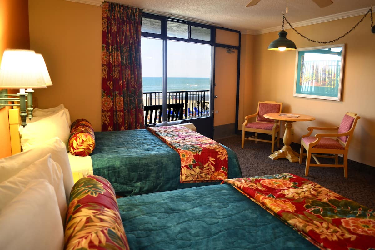 A hotel room with two beds, a small table, two chairs, a view of the beach, and a balcony.