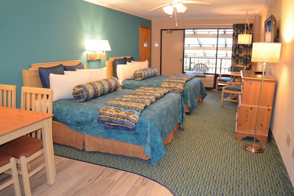 A hotel room with two beds, a wooden desk, chairs, a lamp, and a view of the outdoors.