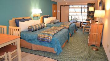 A hotel room with two beds, a wooden desk, chairs, a lamp, and a view of the outdoors.