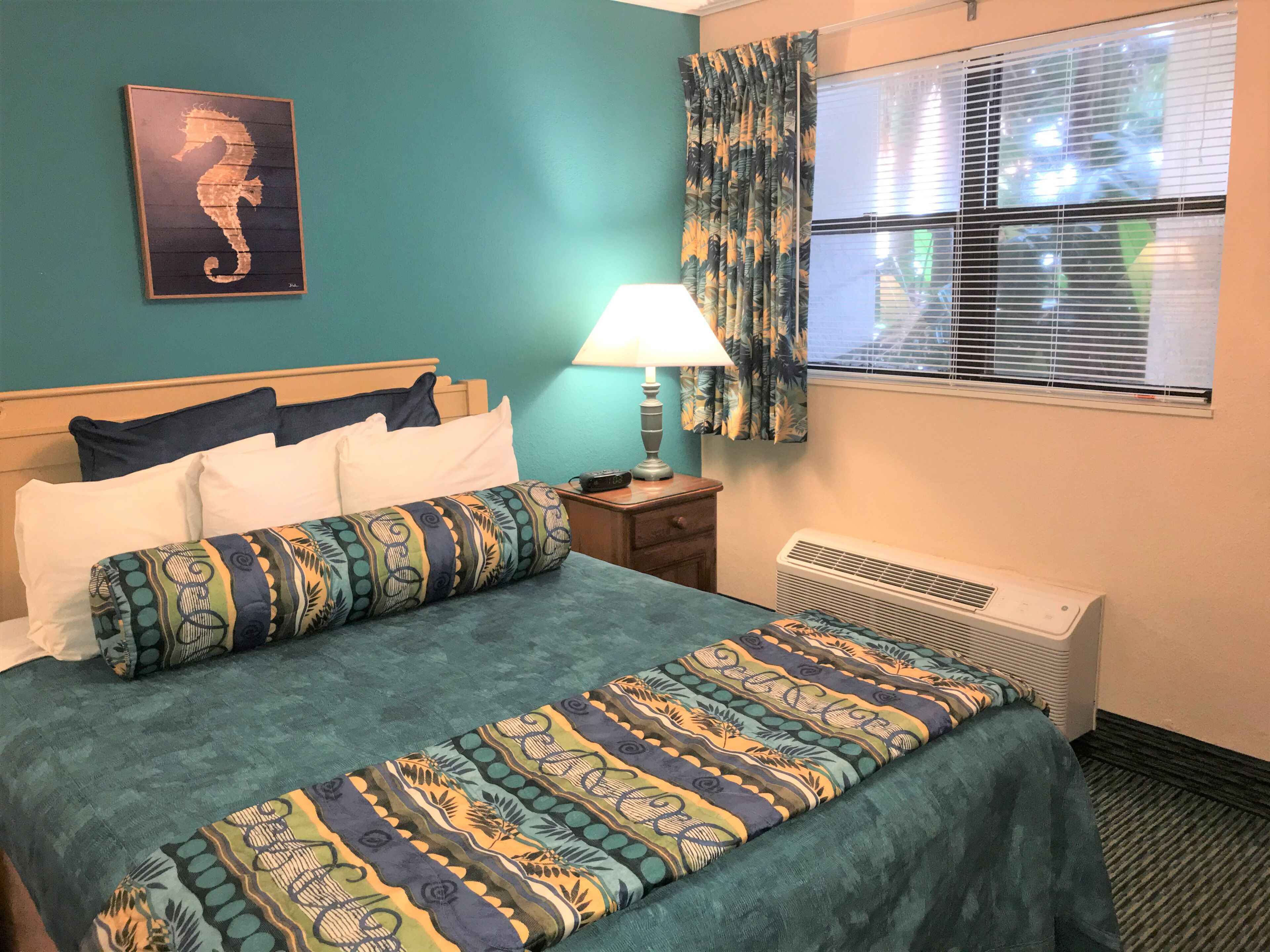 Une chambre d’hôtel comprenant un lit, une table de chevet, une lampe, une fenêtre avec des rideaux et un climatiseur fixé au mur.
