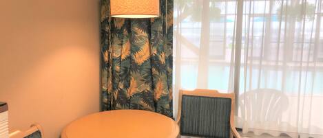 Una habitación con una mesa redonda y dos sillas, una lámpara colgante y vistas a una piscina exterior a través de cortinas translúcidas.