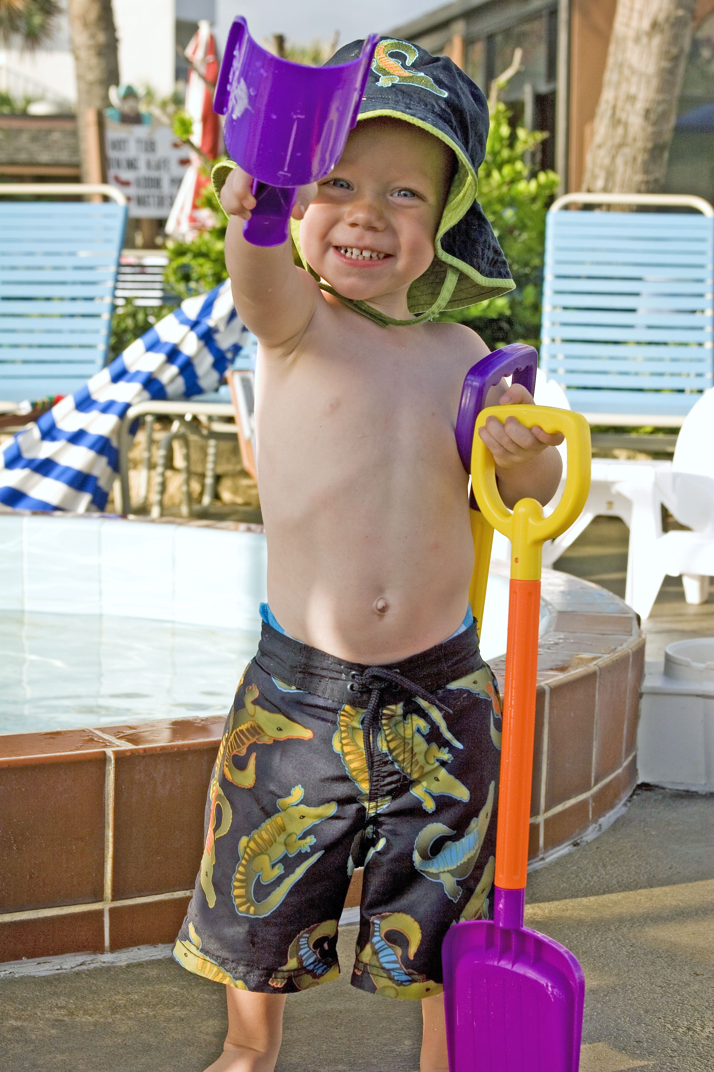 A child holding a purple shovel and a purple bucket, standing near a pool with blue chairs and a white chair in the background.