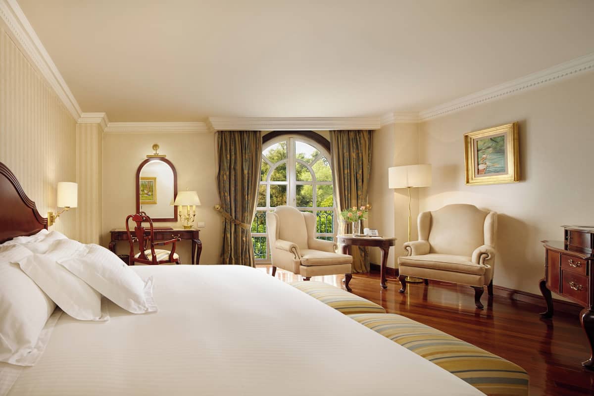 Une chambre d’hôtel avec un grand lit, deux fauteuils, un bureau et une fenêtre avec des rideaux.