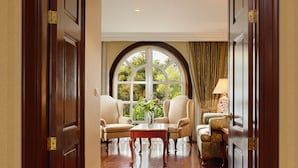 Un pasillo con suelo de madera, un amplio ventanal de arco y vistas al exterior con vegetación. Hay dos sillones y una mesa con un jarrón de flores.