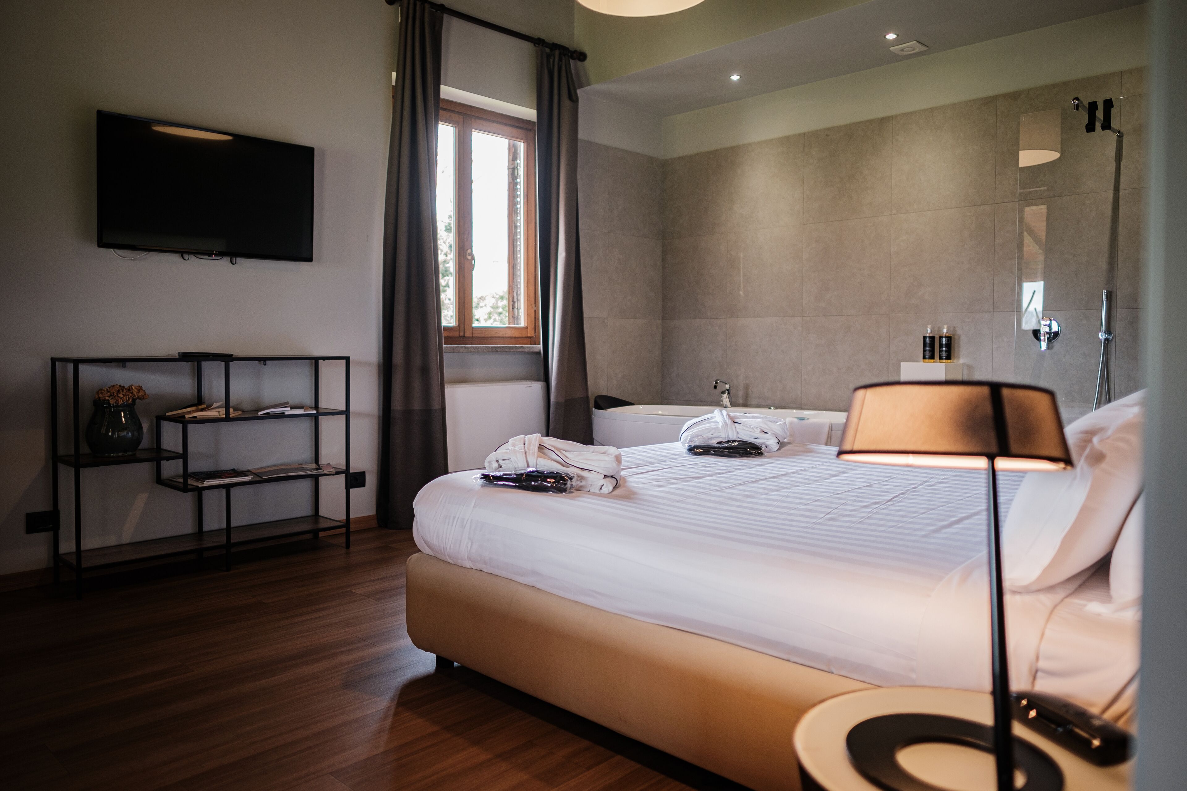 A modern hotel room with a large bed, a bedside table, a TV mounted on the wall, and a window with curtains.