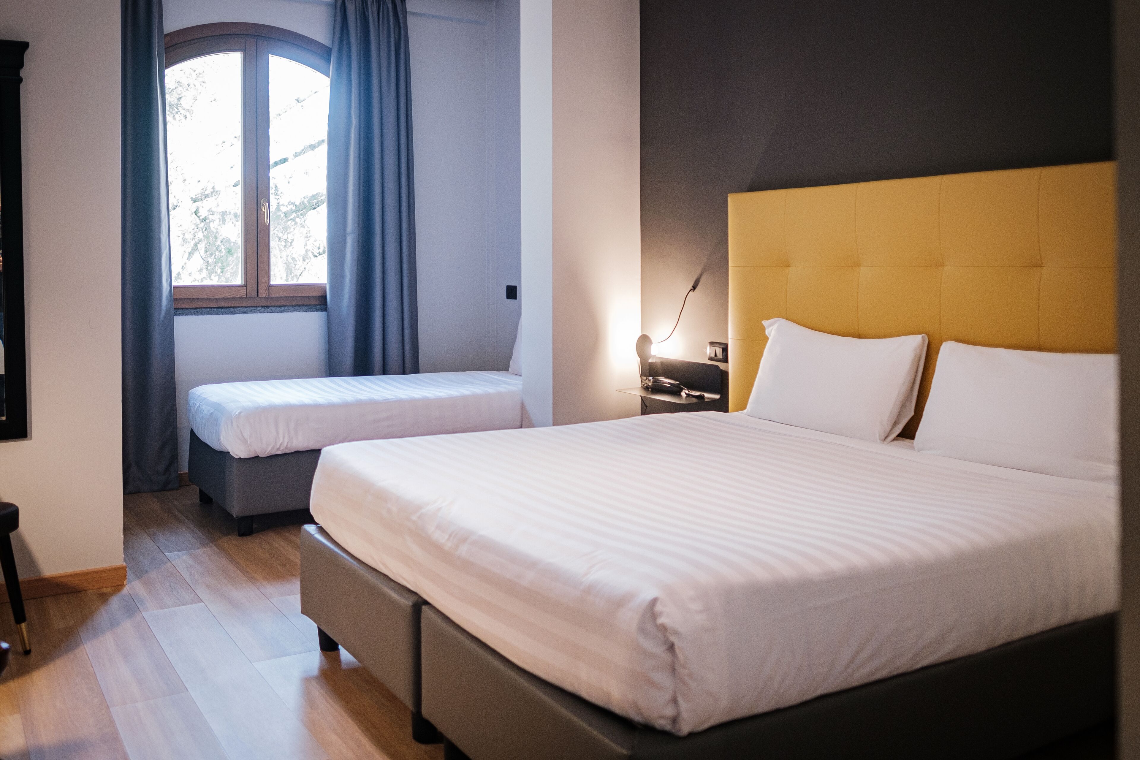 Habitación de hotel con una cama grande, una cama más pequeña, una ventana con cortinas, una mesita de noche y una lámpara.