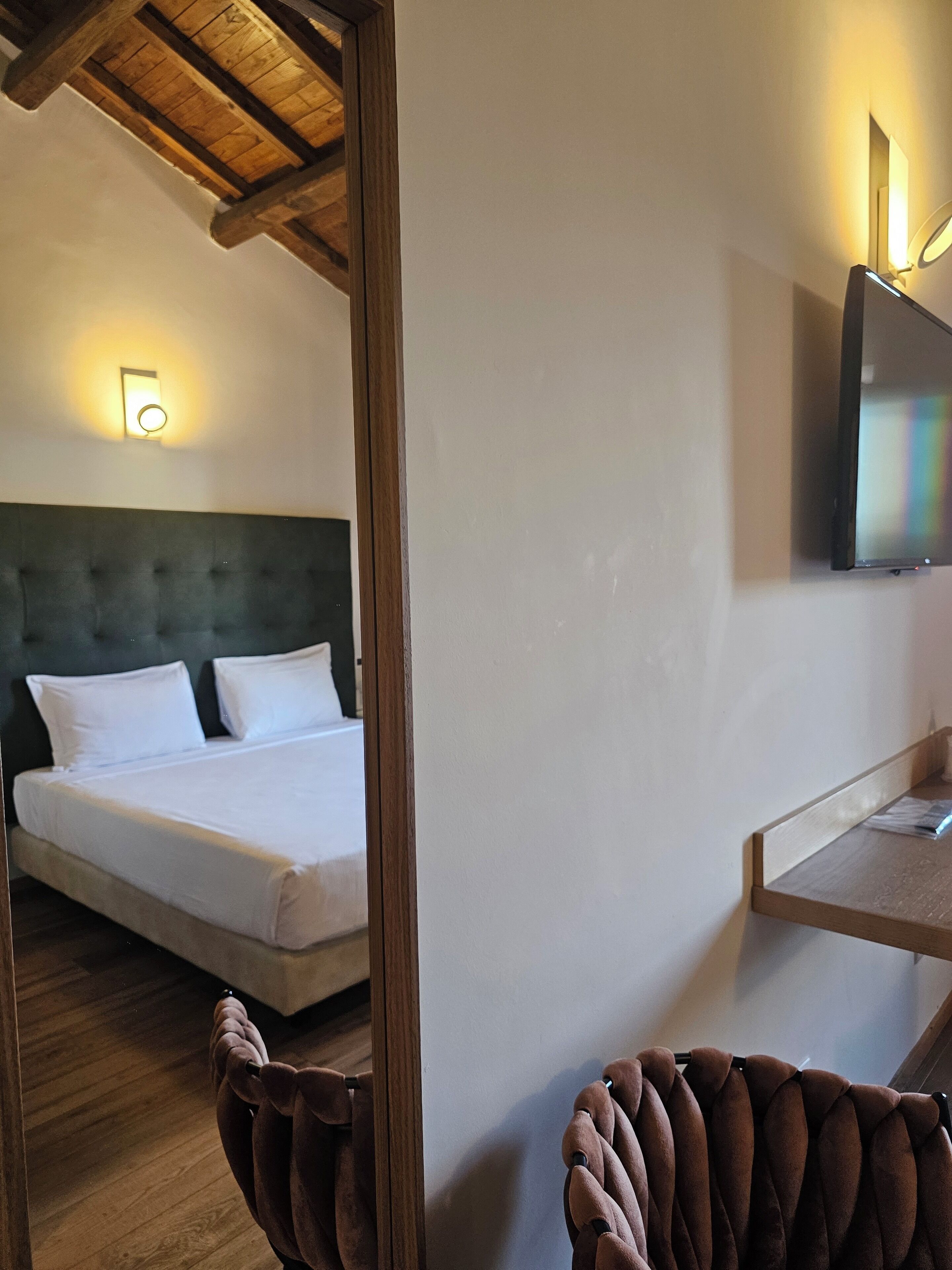 A hotel room with a bed, a television mounted on the wall, and a wooden ceiling.