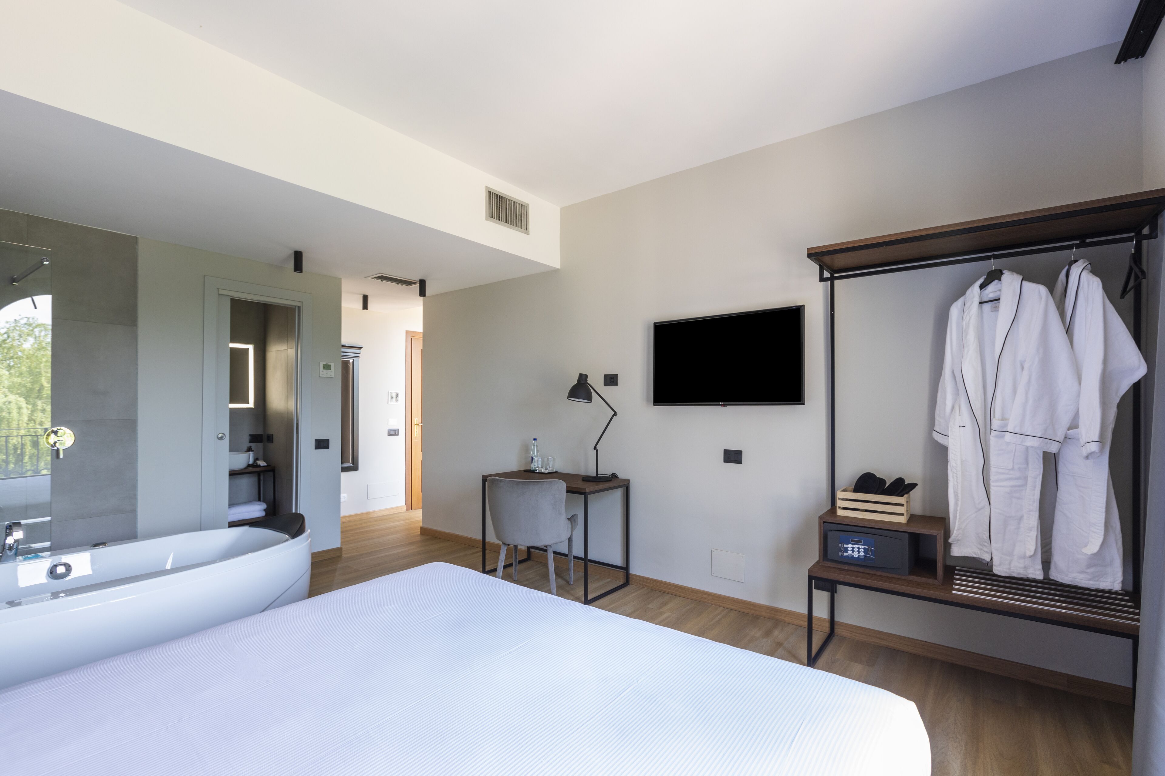 A modern hotel room with a large bed, a bathroom with a bathtub, a desk with a chair, a TV mounted on the wall, and a clothing rack with robes.