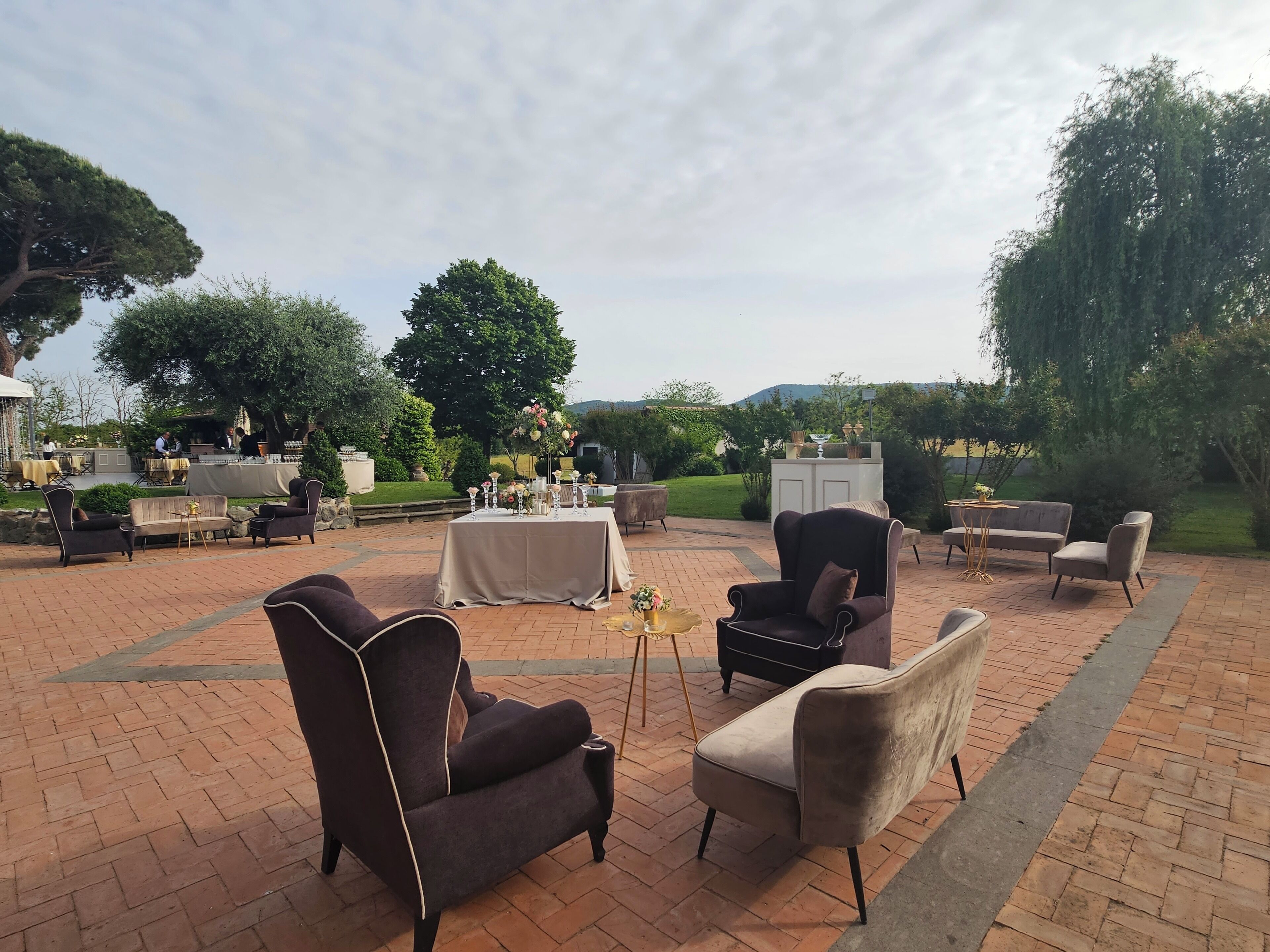 Un patio al aire libre con suelo de ladrillo, varias mesas dispuestas para comer y diferentes opciones de estar, como sillones y sofás.