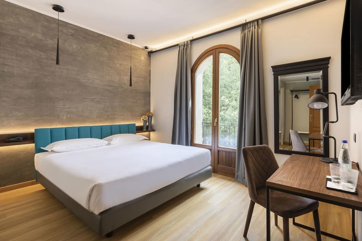 Una habitación de hotel con cama, escritorio, silla, espejo y ventana con cortinas.