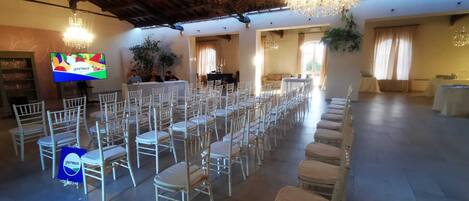 A banquet hall with rows of white chairs, a television screen, and a chandelier.