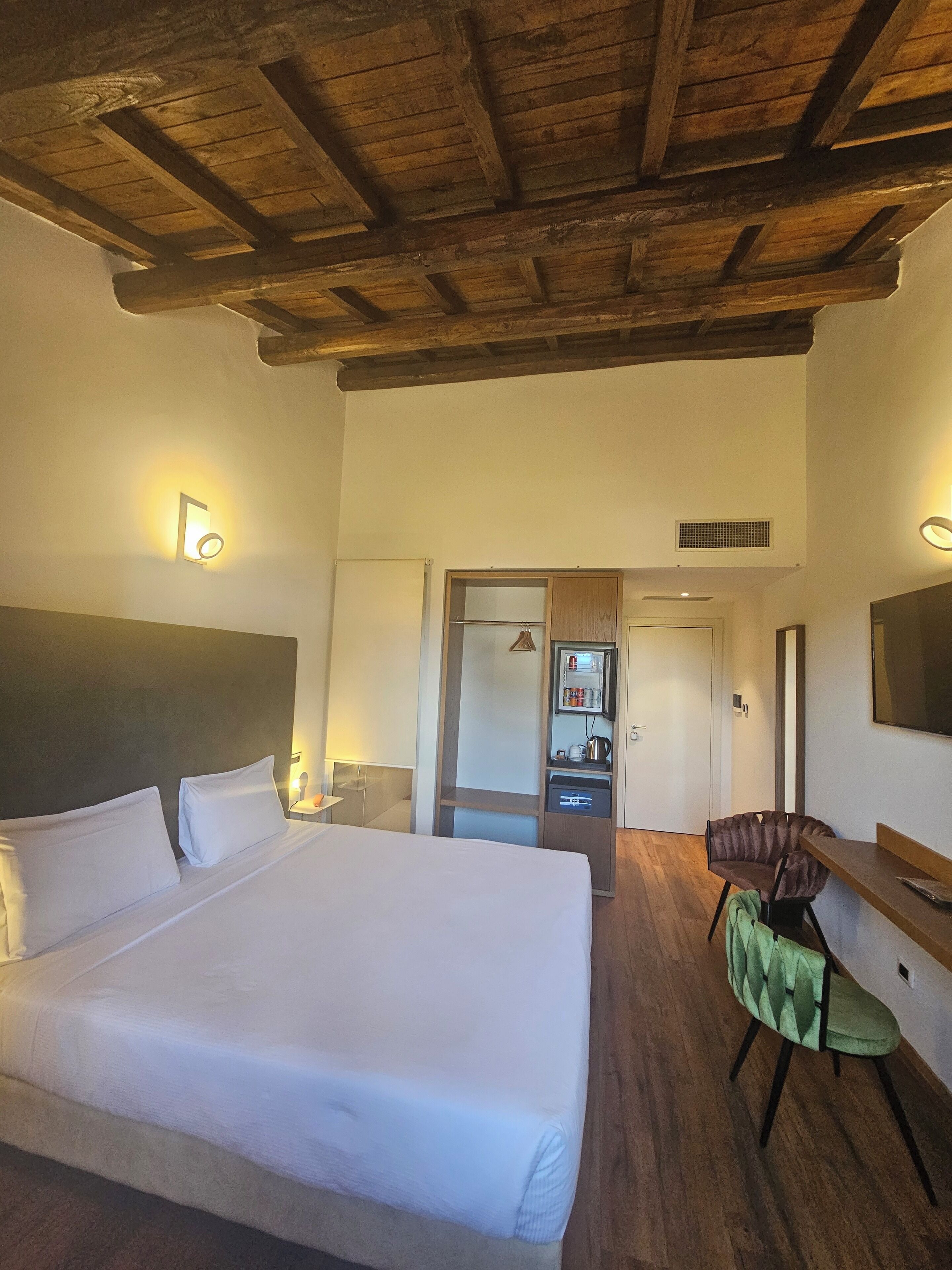 A hotel room with a large bed, a desk with a chair, a wardrobe, and a television on the wall.