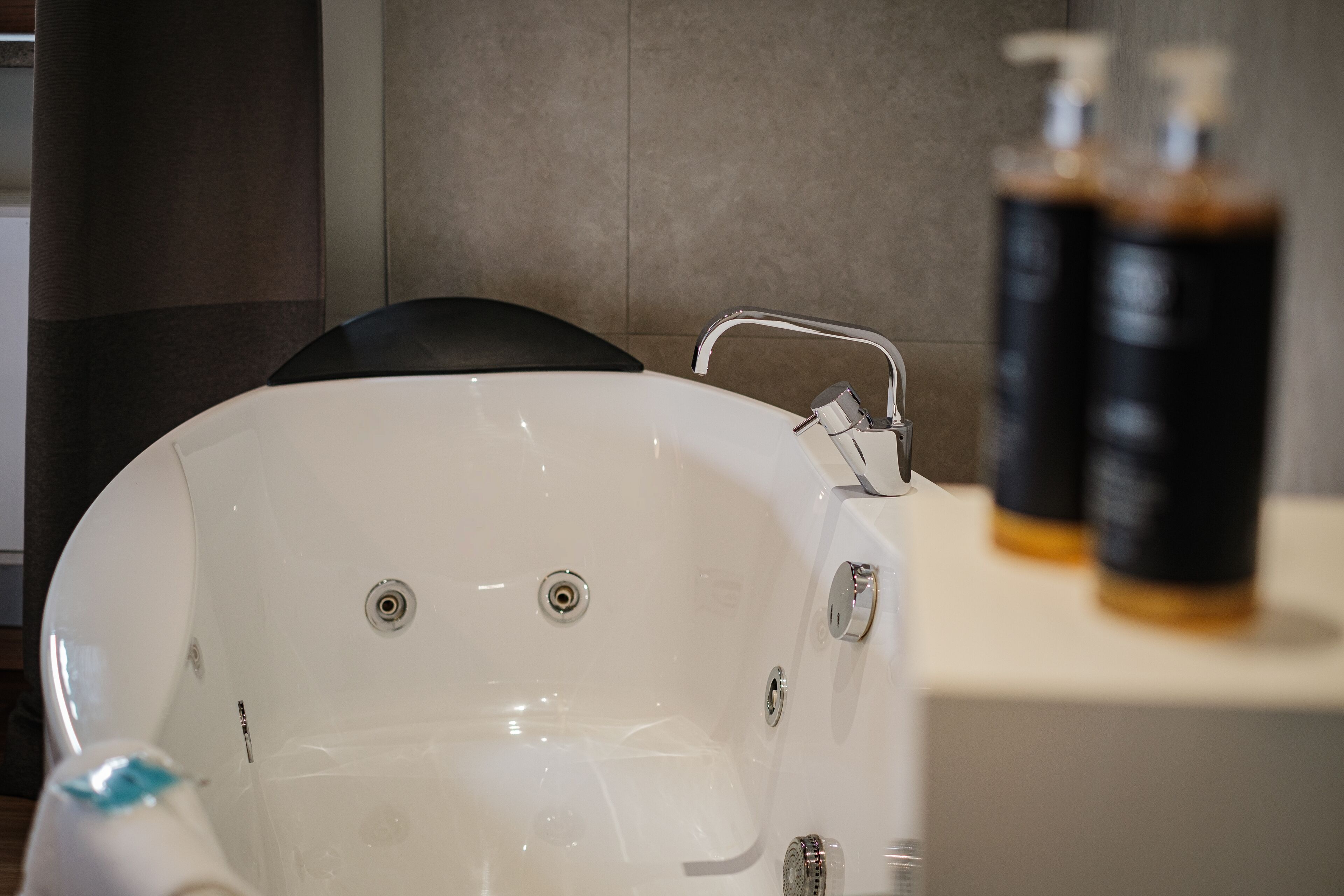 A modern bathroom with a white bathtub, a black chair, and two bottles of shampoo on a shelf.