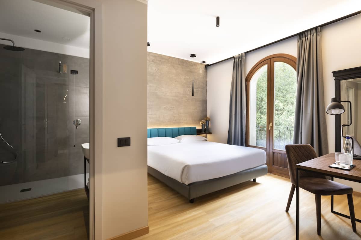 Un dormitorio moderno con una cama grande, un escritorio con silla, ducha y vistas a zonas verdes por una ventana.