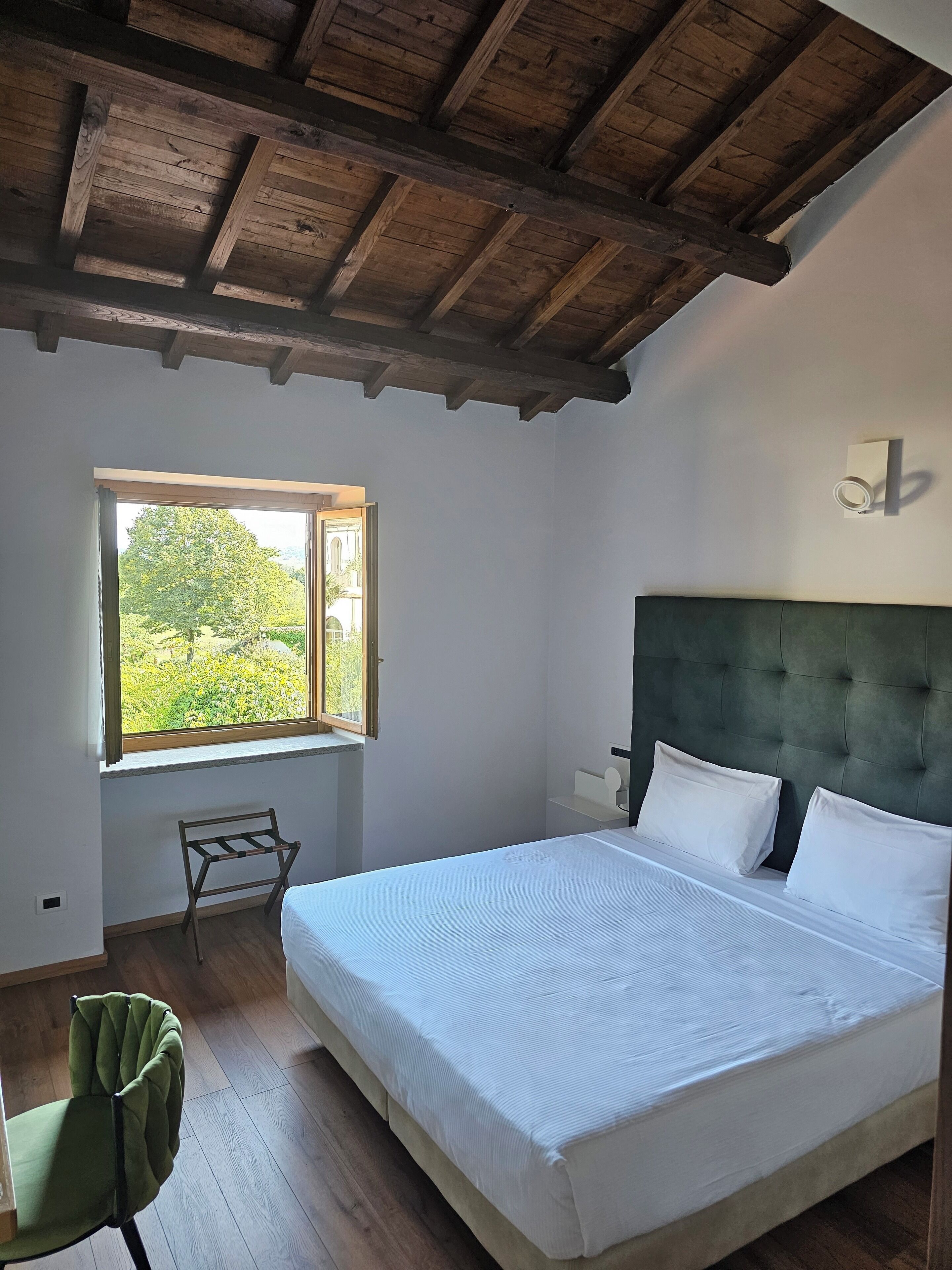 A bedroom with a large bed, a wooden ceiling, a window with a view of greenery, and a green chair.