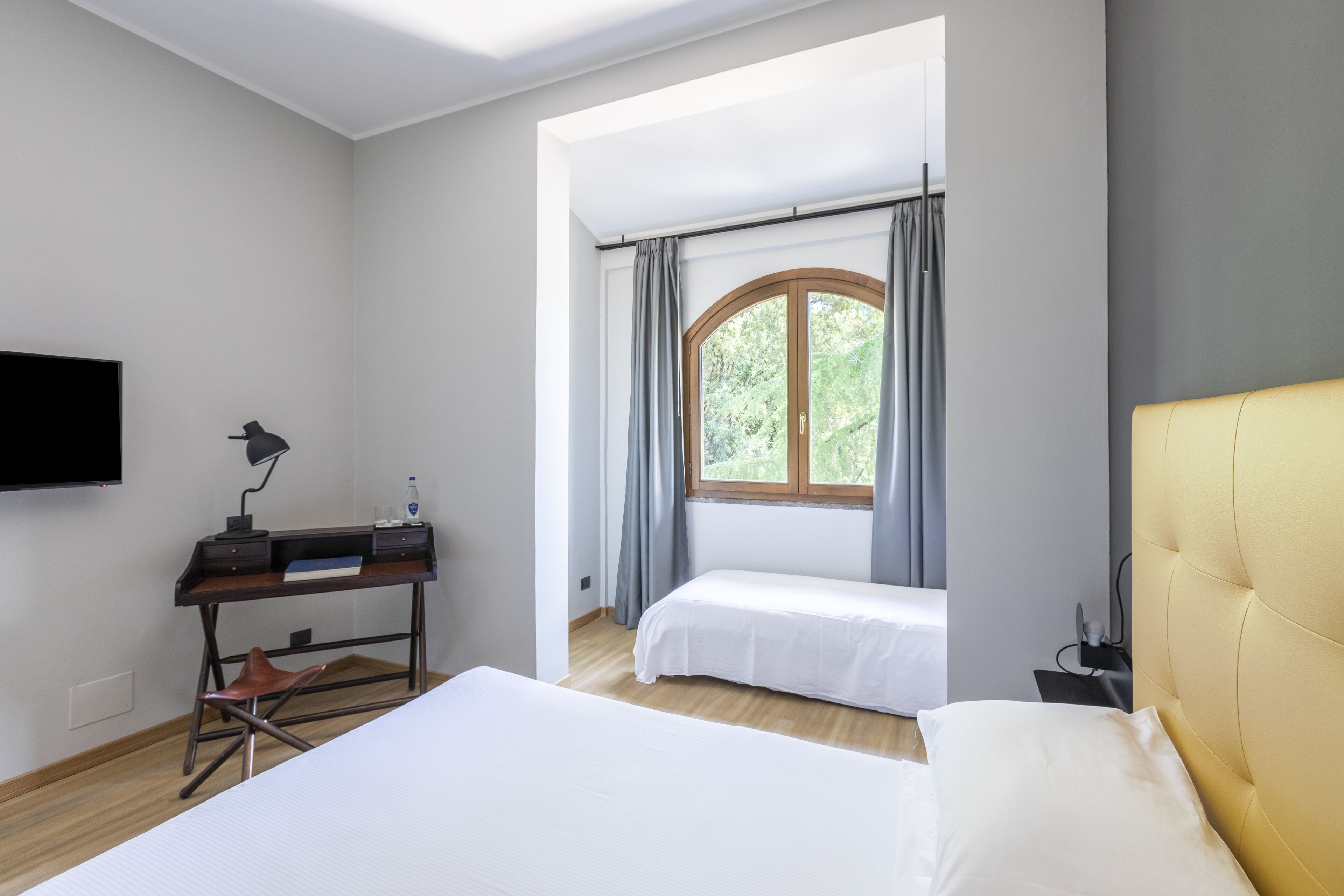 Habitación de hotel con una cama grande, una cama más pequeña, un escritorio con silla, televisor montado en la pared y una ventana con cortinas.