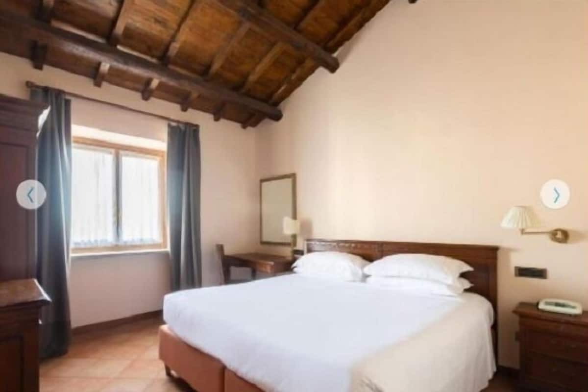 Habitación de hotel con techo de madera, cama con ropa de cama blanca, mesita de noche, ventana con cortinas y un reloj de pared.