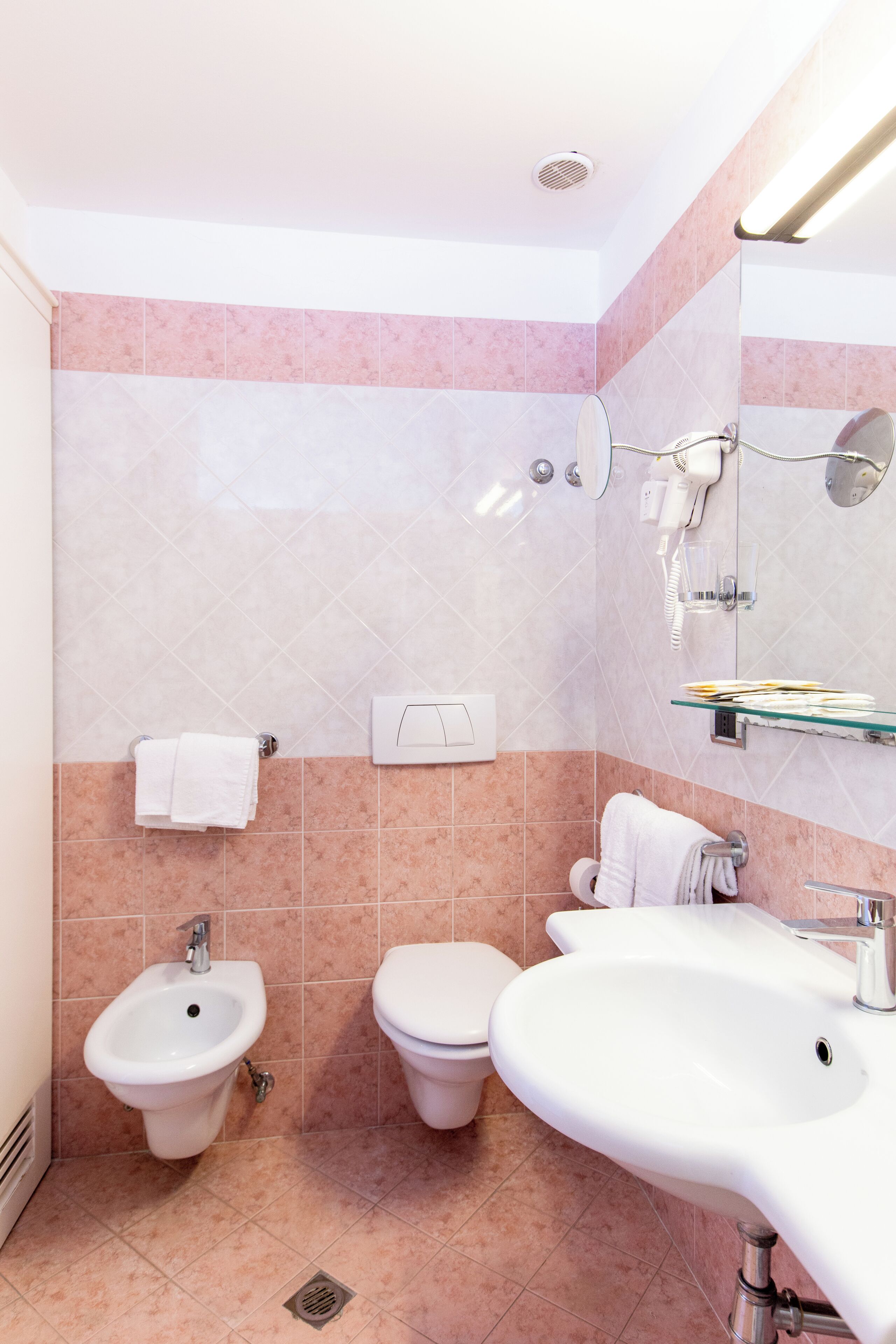A bathroom with a bidet, toilet, and sink, featuring pink and white tiles, a glass shelf, and a hairdryer mounted on the wall.