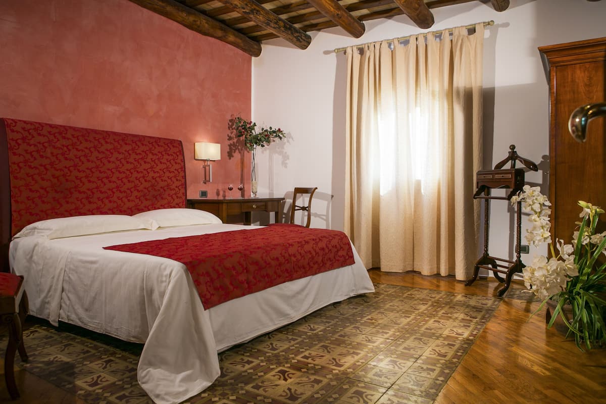 A bedroom with a large bed, a wooden headboard, a desk with a lamp, a chair, a potted plant, and a wooden wardrobe.