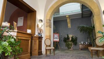 Um hall de entrada com balcão de recepção, uma cadeira de madeira, um quadro em cavaletes e plantas em vasos.