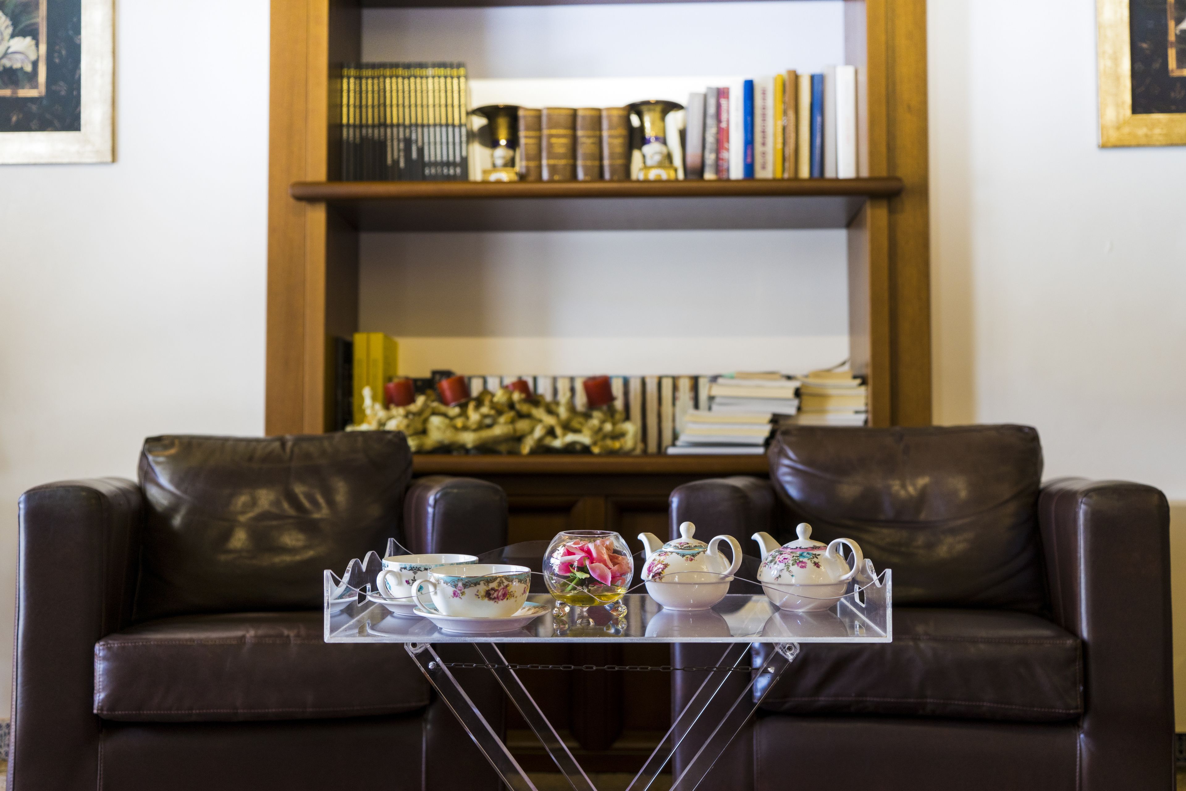 Un sofá de cuero marrón con una mesa de centro de vidrio al frente. Detrás del sofá, hay una estantería de madera con libros y objetos decorativos.