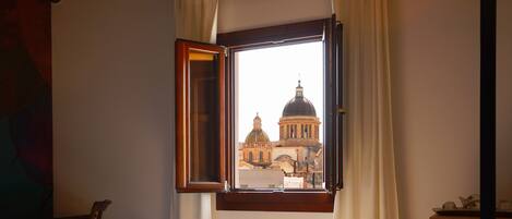 A room with a bed, a window with a view of a building, a wooden ceiling, and a patterned floor.