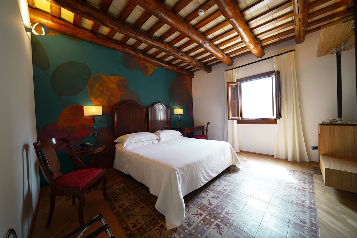 A bedroom with a bed, wooden headboard, a chair, a desk, and a window with curtains.