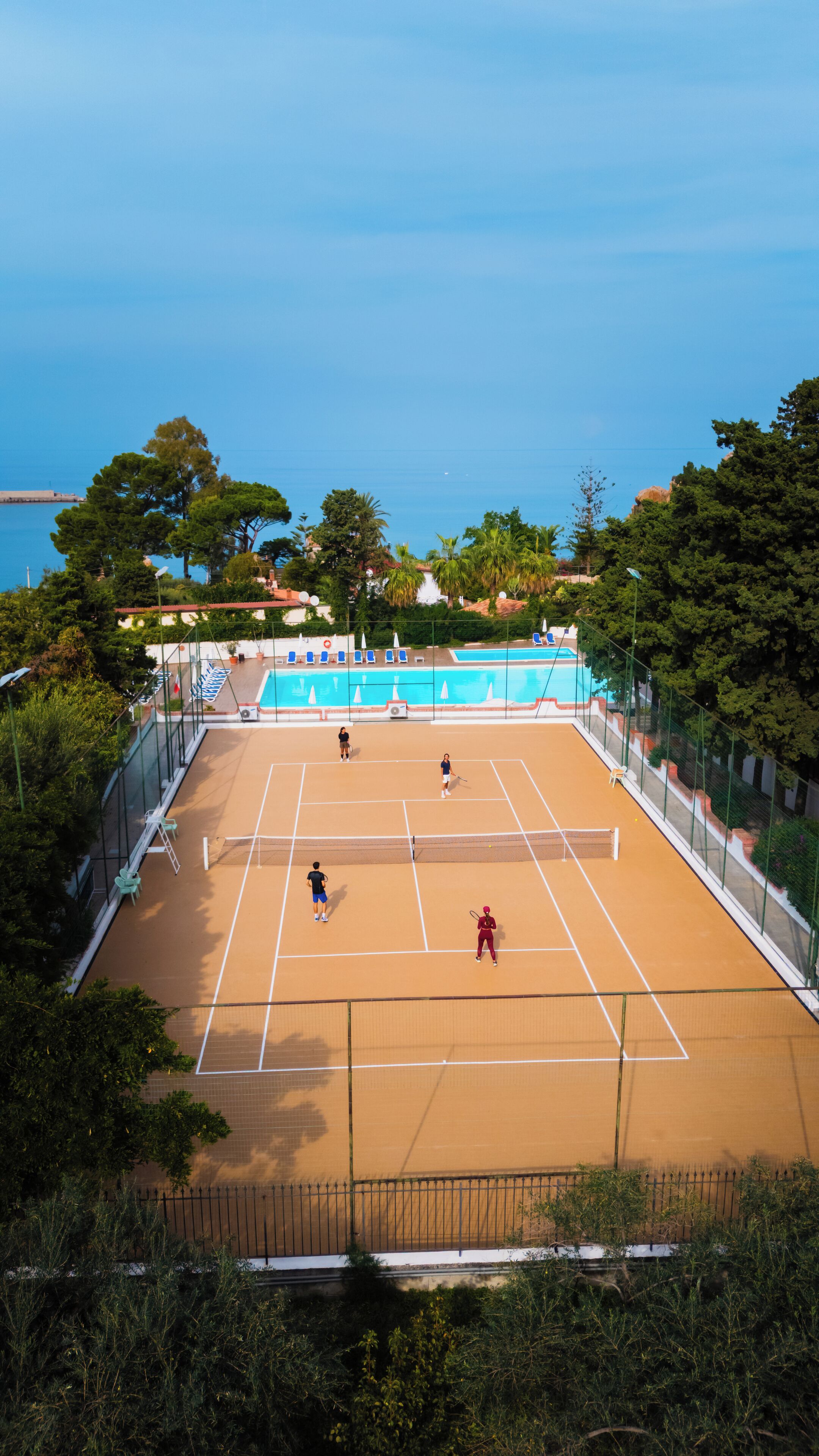 Ein Tennisplatz im Freien mit Netz, umgeben von Bäumen und einem Pool im Hintergrund.