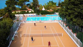 Ein Tennisplatz im Freien mit Netz, umgeben von Bäumen und einem Pool im Hintergrund.