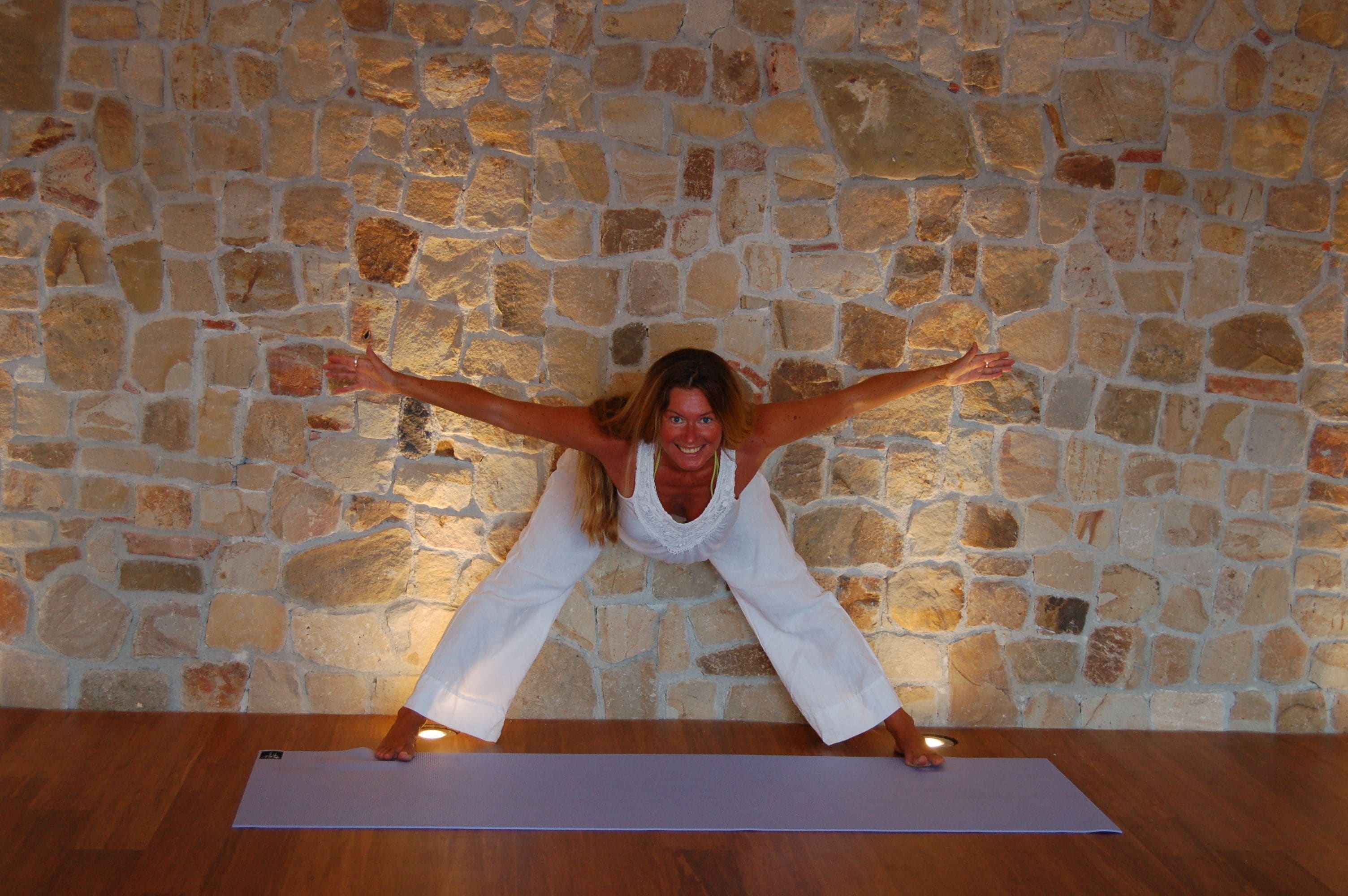 A person in a white outfit performing a yoga pose on a mat against a stone wall.