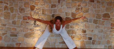 A person in a white outfit performing a yoga pose on a mat against a stone wall.