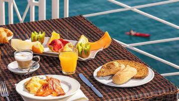Una mesa de desayuno con cruasanes, huevos, tocino, tostadas, fruta y jugo, sobre una mesa de mimbre junto a una barandilla con vista al mar.