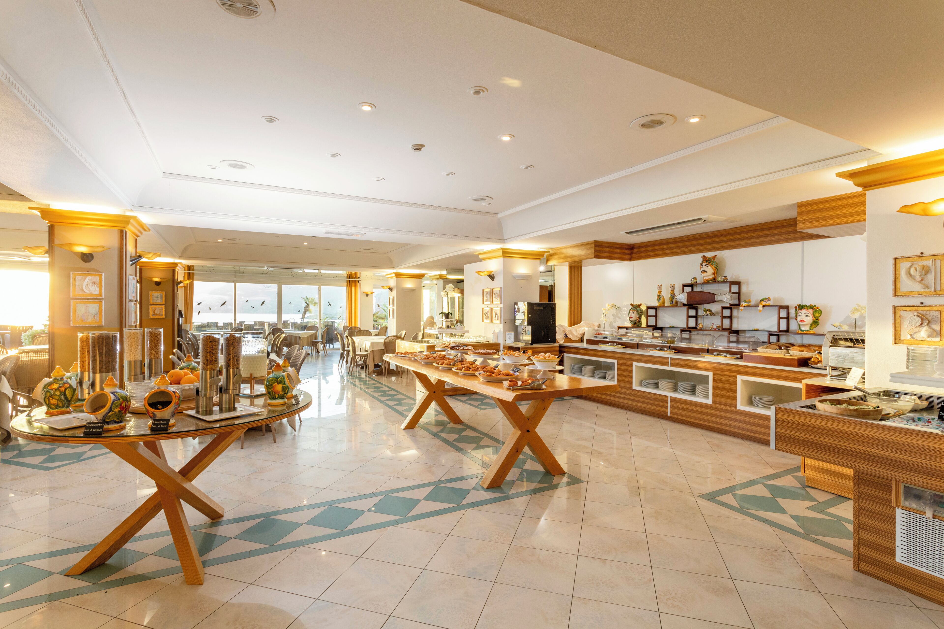 Spaziosa sala da pranzo dell'hotel con diverse postazioni del buffet, un tavolo centrale per il servizio e vista sull'area salotto esterna.