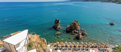 Un hotel frente al mar con aguas azul cristalino, afloramientos rocosos y una zona de playa con tumbonas y sombrillas.