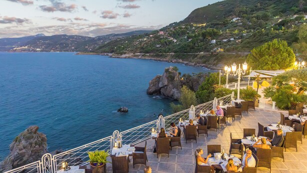 A coastal restaurant with outdoor seating overlooking the sea, featuring wicker furniture and a scenic view of the coastline. - Hotel Kalura (Cefalù)